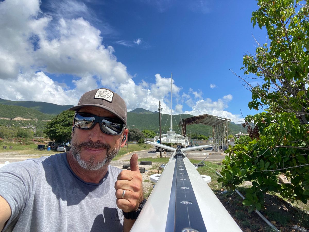 Collin Linehan giving a thumbs up beside an unstepped catamaran mast in the boatyard